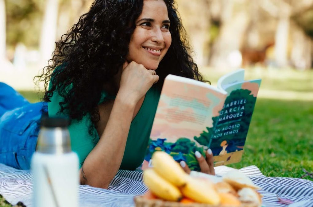 Smiling woman reading a book at a picnic in a sunny park setting.