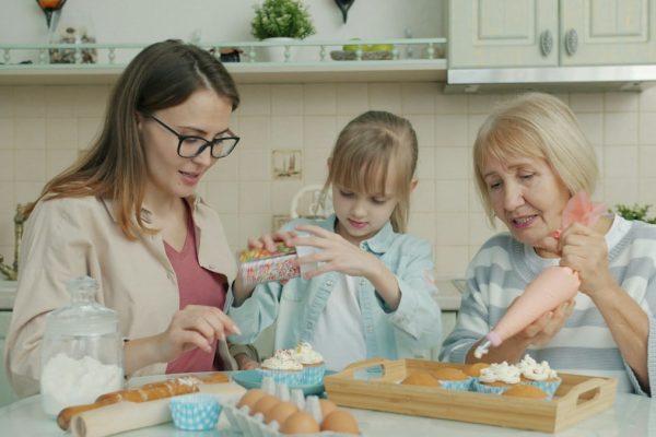Happy family kid, young mom and senior granny are decorating cupcakes busy with culinary activity in kitchen. Apartment, people and cookery concept.